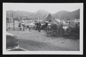 Group of people in front of a tent, cars, and buildings with desert mountains in background: photographic print