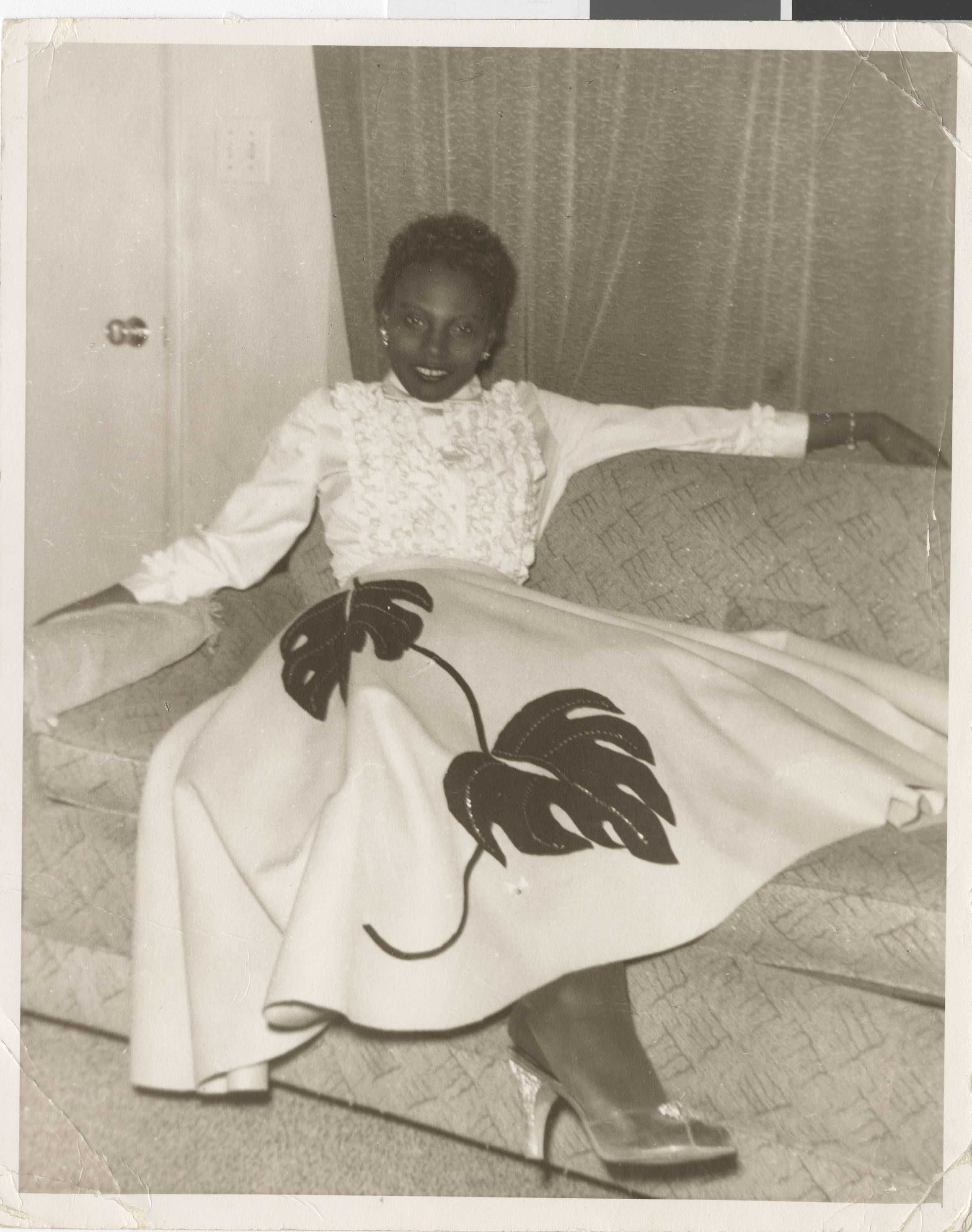 Black and white photograph of Ruby Amie-Pilot seated on a sofa in a long skirt, no date